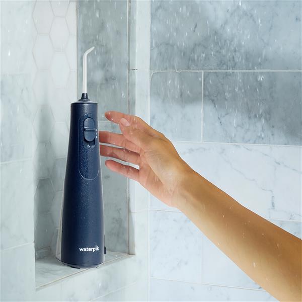 Hydropulseur sans fil 1000 bleu sur un rebord de douche, une main tendue vers l’appareil.